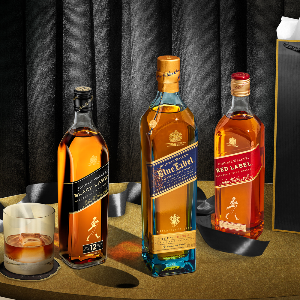 Three bottles of Johnnie Walker whisky—Black Label, Blue Label, and Red Label—are displayed on a table next to a glass with whisky and ice, with a gift bag in the background.