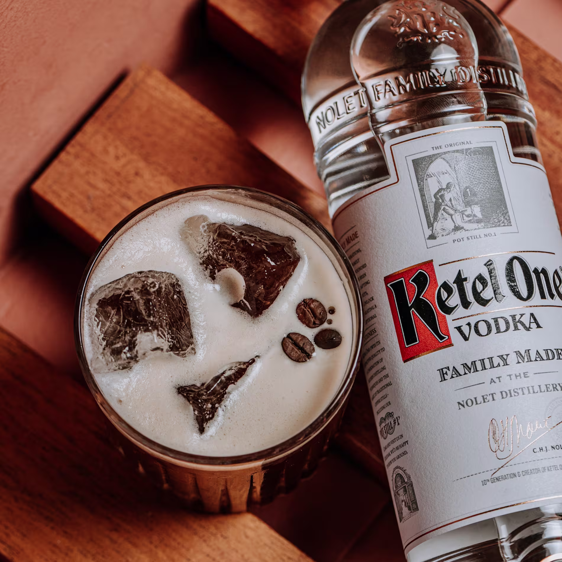 Un cóctel con espuma y granos de café flota entre hielos, junto a una botella de Ketel One sobre una superficie de madera cálida.