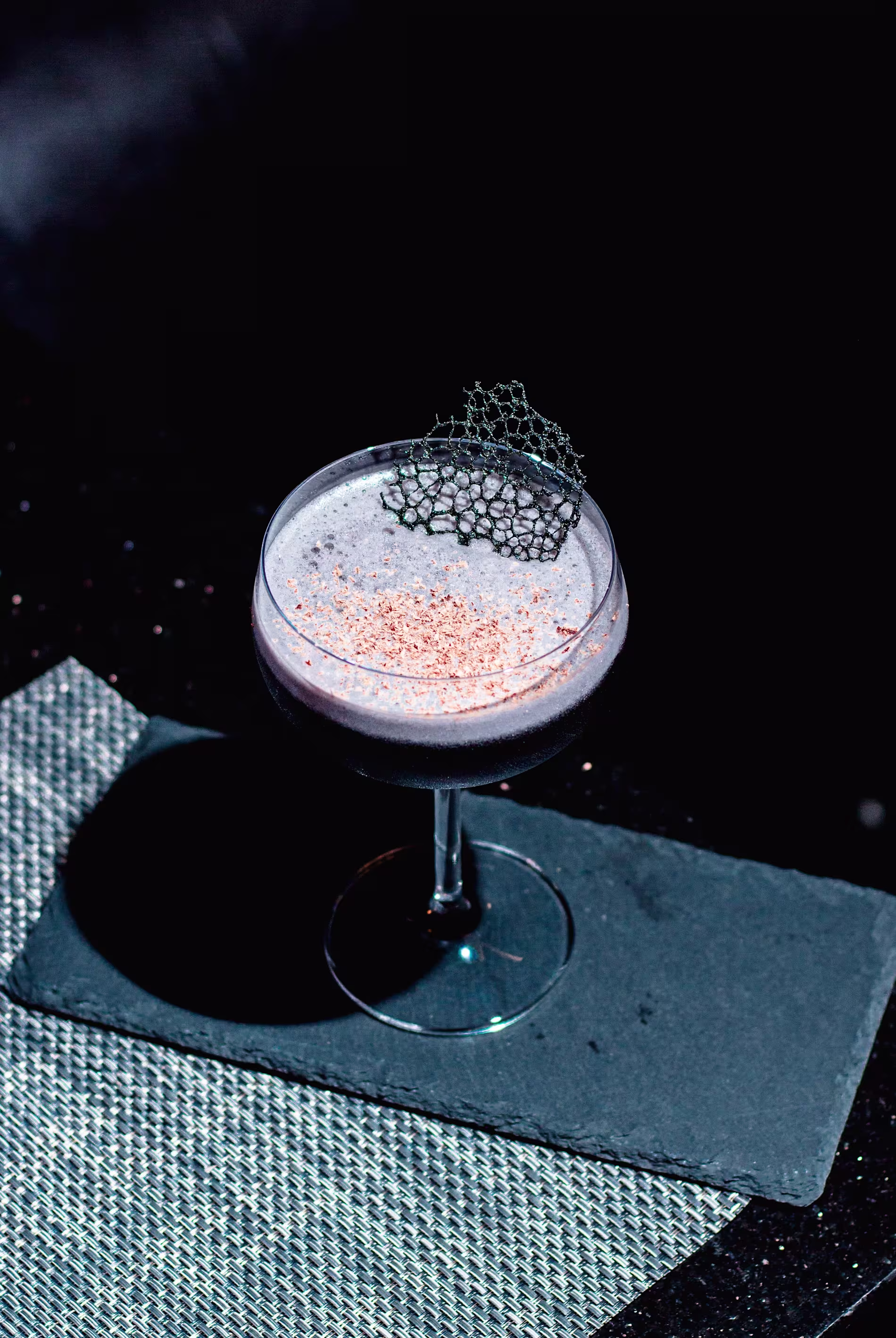 A dark cocktail in a coupe glass topped with a thin, decorative garnish, placed on a black rectangular slate and a woven placemat.