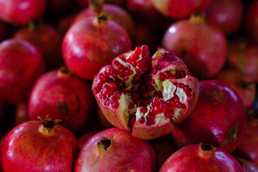 A pile of whole pomegranates with one pomegranate in the center split open, exposing its red seeds.