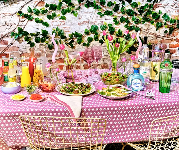 A table with a pink-patterned cloth set with salads, drinks, liquor bottles, glassware, flowers, and food, against a brick wall with green vines.