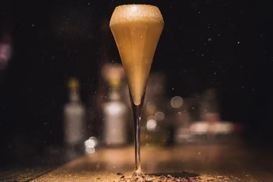 A tall champagne flute filled with a frothy golden beverage sits on a dimly lit surface, with bubbles forming at the top and a blurred background.