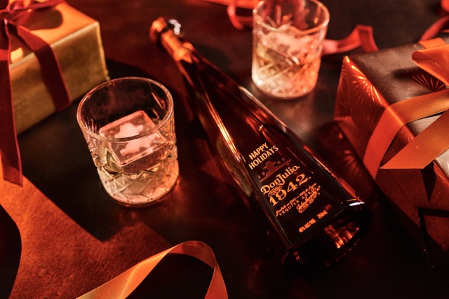A bottle of Don Julio 1942 tequila lies on a table surrounded by two glasses with ice and wrapped holiday gifts with red ribbons.
