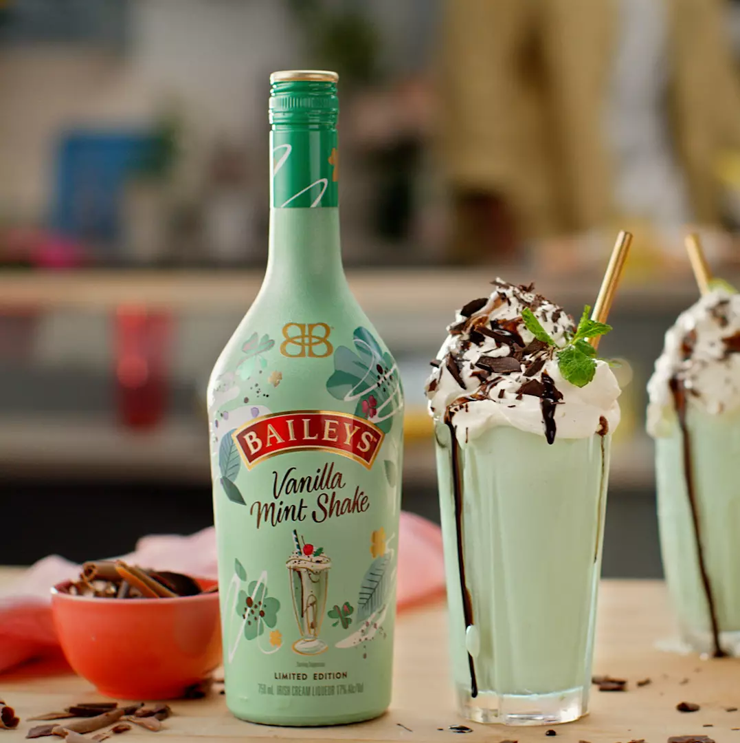 A bottle of Baileys Vanilla Mint Shake stands next to a glass of mint green milkshake topped with whipped cream, chocolate shavings, and a mint sprig.