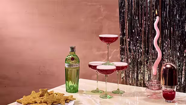 A bottle of gin, a tower of four coupe glasses with red cocktails, star-shaped cookies, and a pink twisted candle on a marble surface with a pink and silver backdrop.