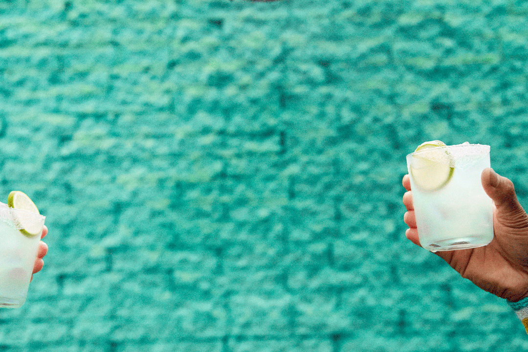 Two hands hold glasses with salted rims, filled with a light-colored drink and garnished with lime wedges, in front of a turquoise textured wall.