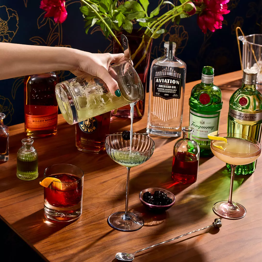 A hand pours a green cocktail into a glass on a wooden table with assorted bottles of liquor, garnished drinks, and bar tools.