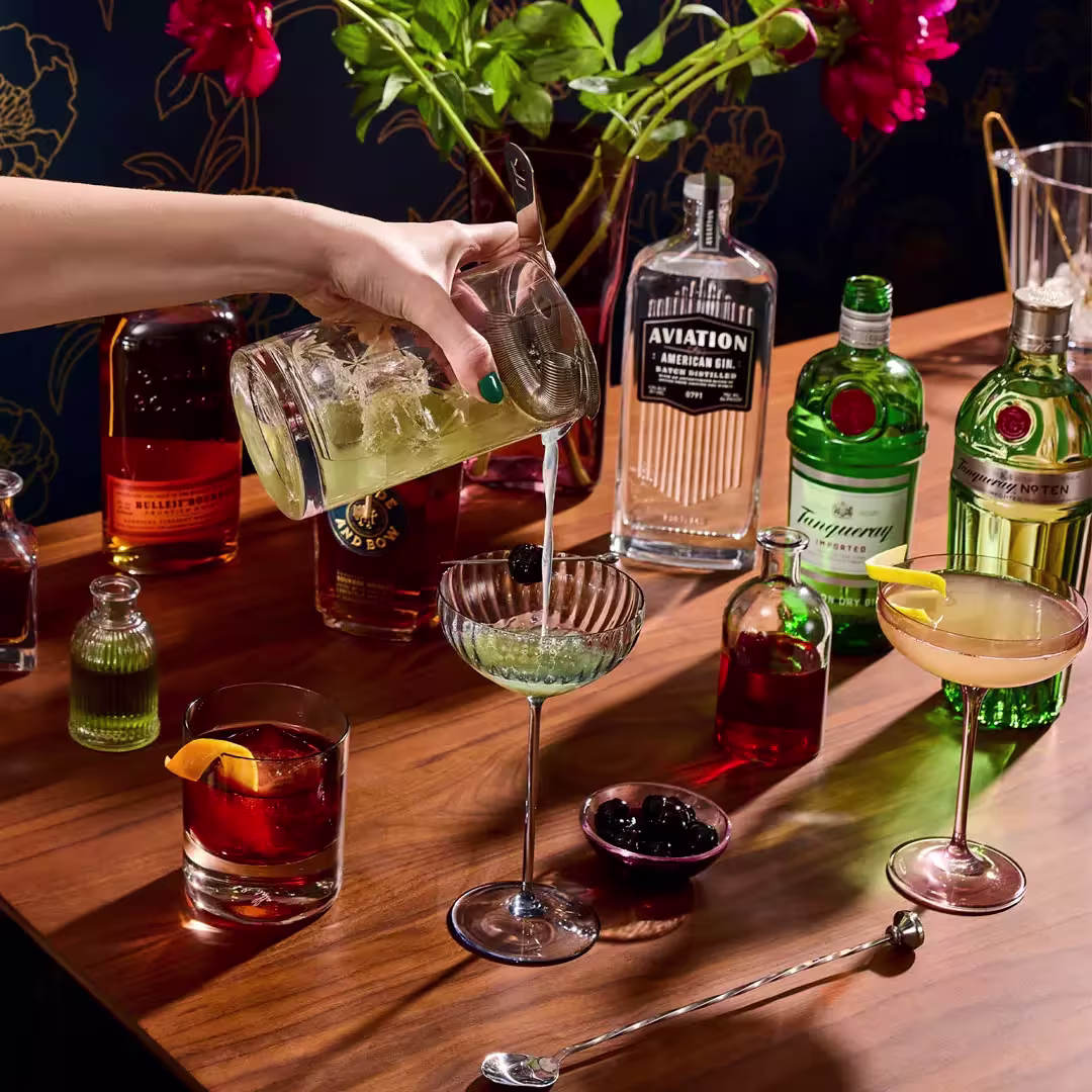A hand pours a green cocktail into a glass on a wooden table with assorted bottles of liquor, garnished drinks, and bar tools.