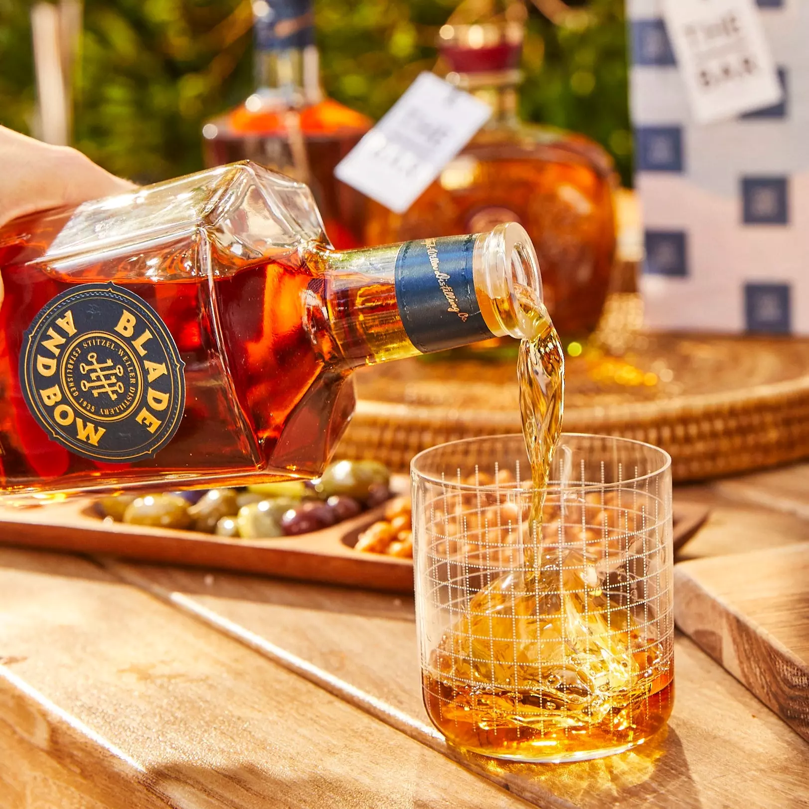 A bottle of Blade and Bow whiskey is being poured into a glass with ice on a wooden table, with snacks and other bottles in the background.