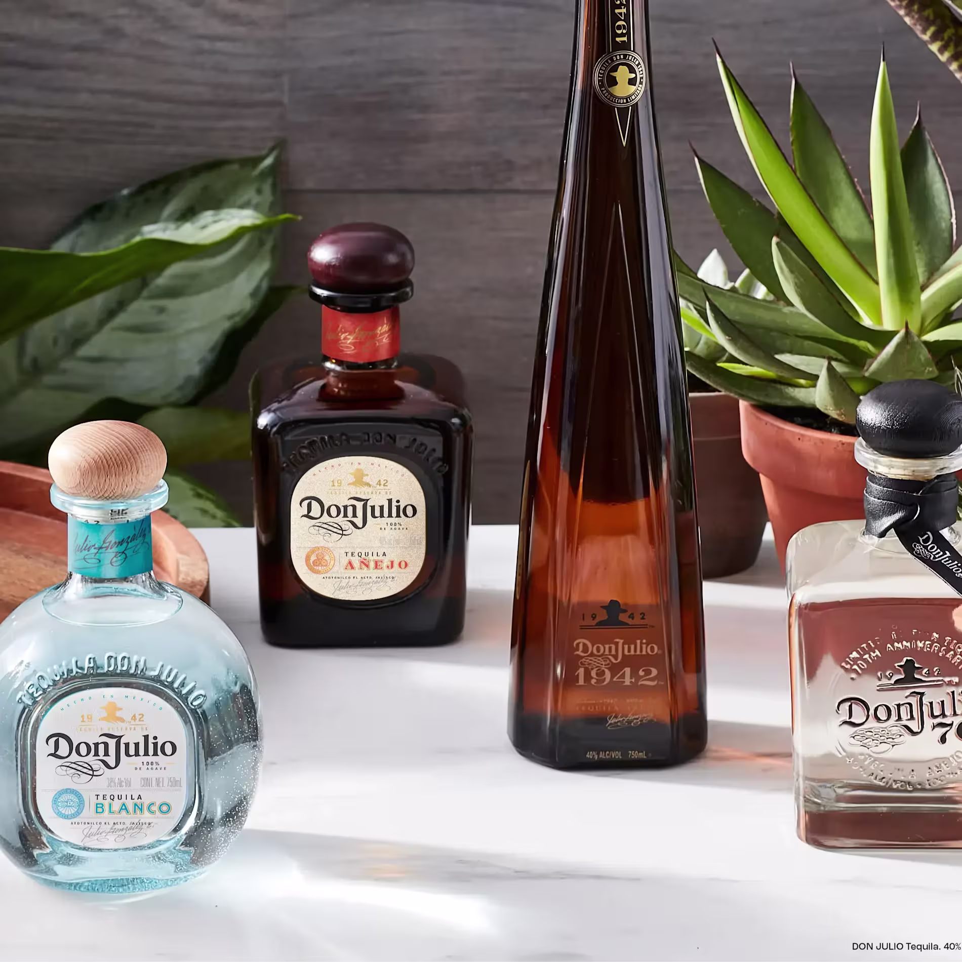 Cuatro botellas de tequila Don Julio -Blanco, Añejo, 1942 y Reposado- se exhiben sobre una superficie blanca con plantas en macetas al fondo.