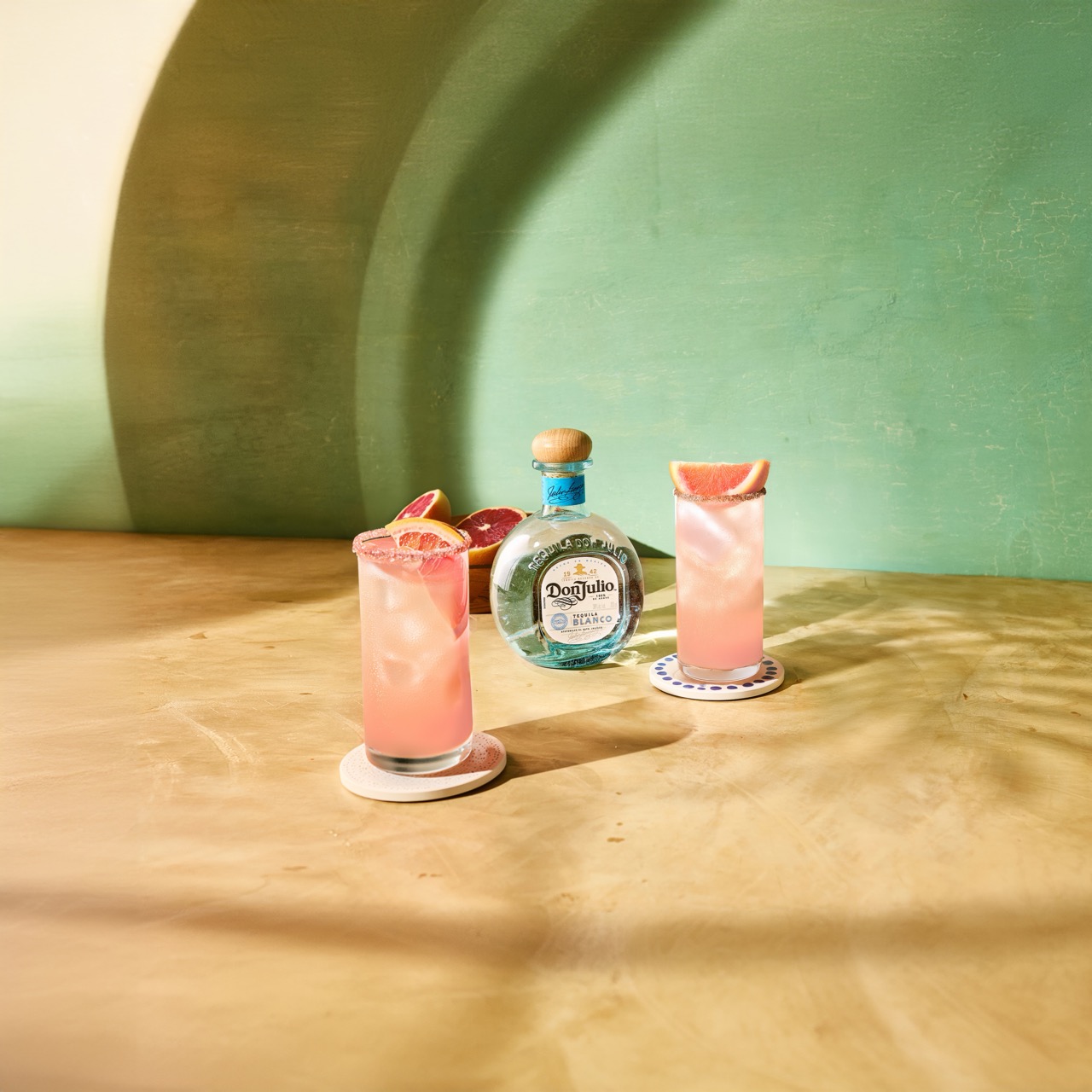 A bottle of Don Julio tequila and two pink cocktails garnished with grapefruit slices are displayed on a wooden surface against a green and beige background.