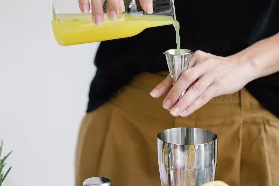 A person pours yellow liquid from a glass bottle into a jigger, preparing to mix a drink with a metal cocktail shaker on the table.