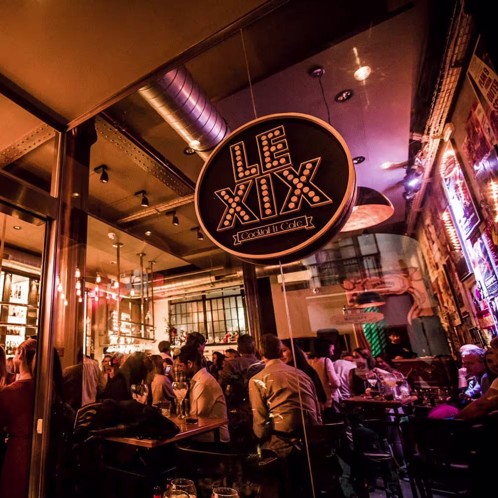 Vista desde el exterior de Le XIX con su cartel luminoso, interior repleto de clientes con copas y ambiente cálido bajo luces colgantes.