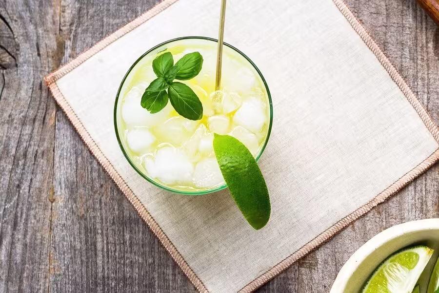 A glass of iced yellow drink garnished with a lime wedge and basil leaves on a cloth napkin, set on a wooden surface.
