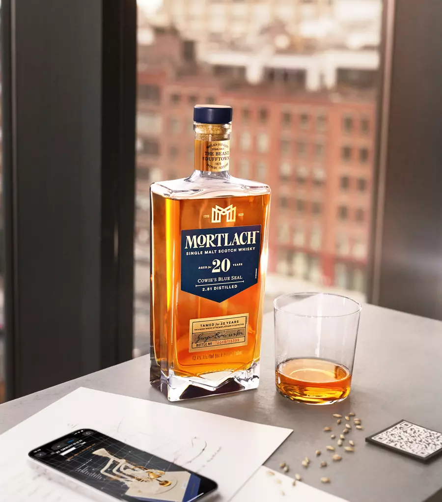 A bottle of Mortlach 20 Year Old single malt scotch whisky stands next to a glass of whisky, papers, and a smartphone on a table by a window with a city view.