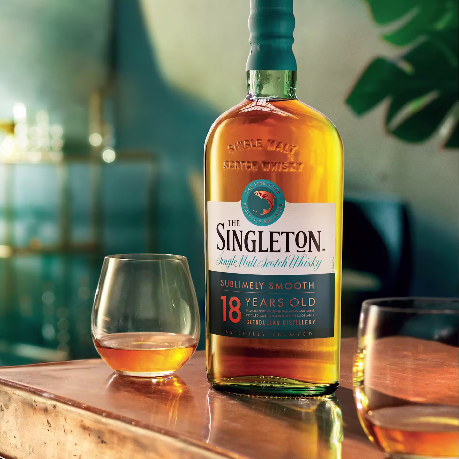 A bottle of The Singleton 18 Years Old single malt scotch whisky sits on a wooden surface beside two whisky glasses, one filled with whisky.