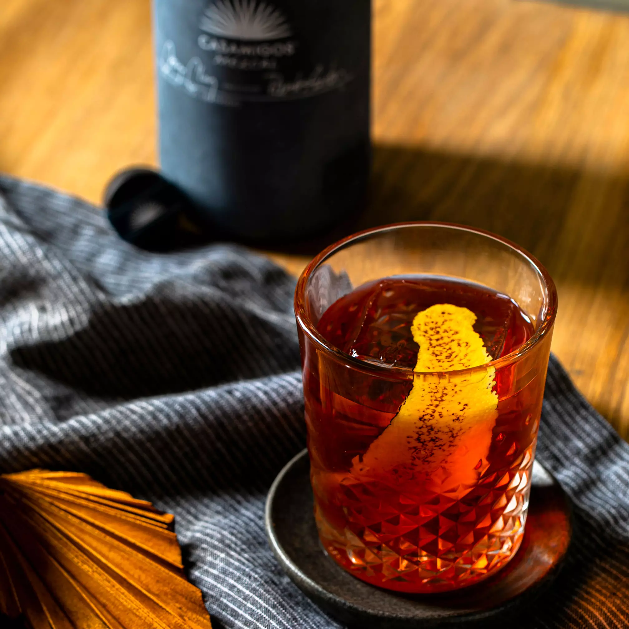 A glass of amber cocktail with a citrus peel garnish sits on a small plate beside a striped cloth and a dark bottle in the background on a wooden surface.