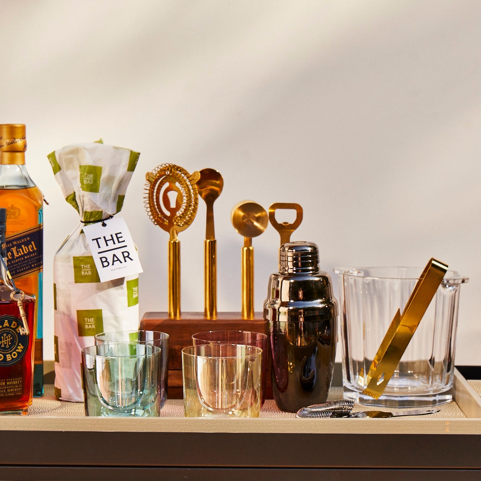 A bar set with bottles, wrapped gift, cocktail tools, shaker, ice bucket with tongs, and two glasses arranged on a tray.
