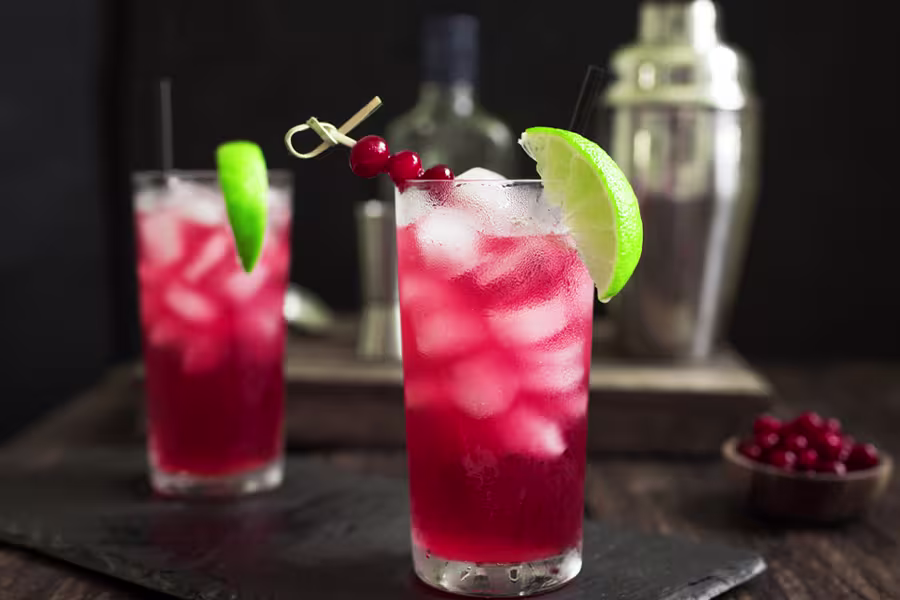 A glass of red cocktail with ice, garnished with a lime wedge and red berries, sits on a table; another cocktail and bar tools are in the background.