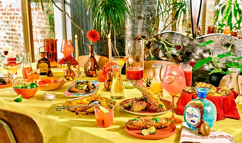 A colorful table is set with grilled corn, various dishes, drinks, limes, and a bottle of tequila, surrounded by plants and bright decor in a sunlit room.