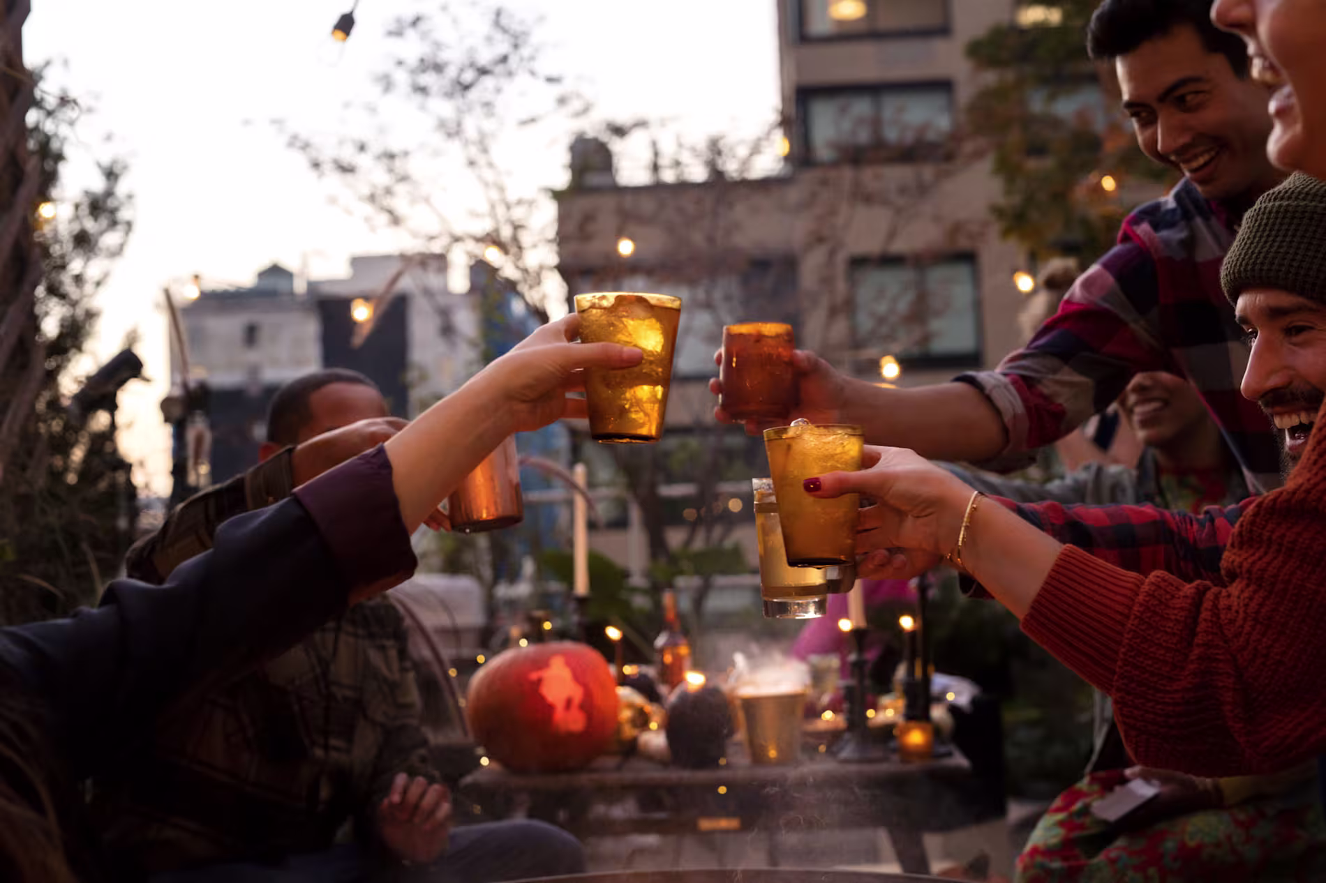A group of people sitting outdoors clink glasses together in a toast, with autumn decorations and string lights visible in the background.