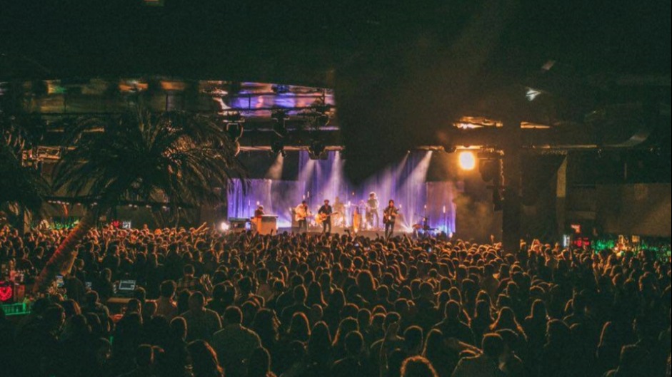 La foto muestra un concierto en vivo con una banda actuando en el escenario en La riviera. El escenario está iluminado por luces de escenario, y hay una gran multitud de espectadores viendo la actuación.