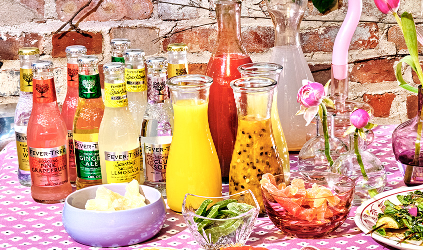 Assorted Fever-Tree mixers, carafes of juice, citrus slices, mint, and flowers are arranged on a pink patterned tablecloth against a brick wall background.