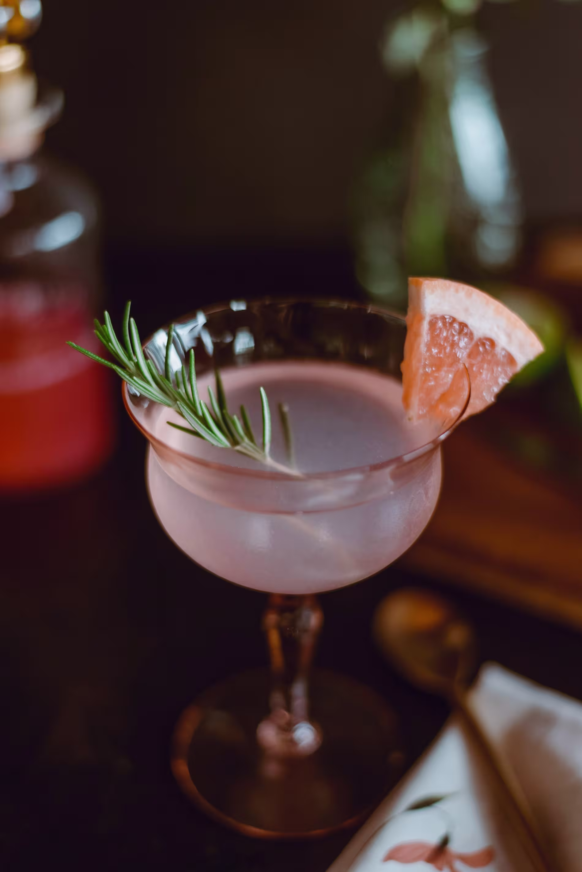 A pink cocktail in a coupe glass garnished with a rosemary sprig and a grapefruit wedge, placed on a dark surface with a spoon and napkin nearby.