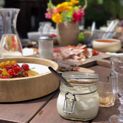 A jar of white sauce with a knife on a wooden table surrounded by glasses, plates of food, and a vase of flowers.