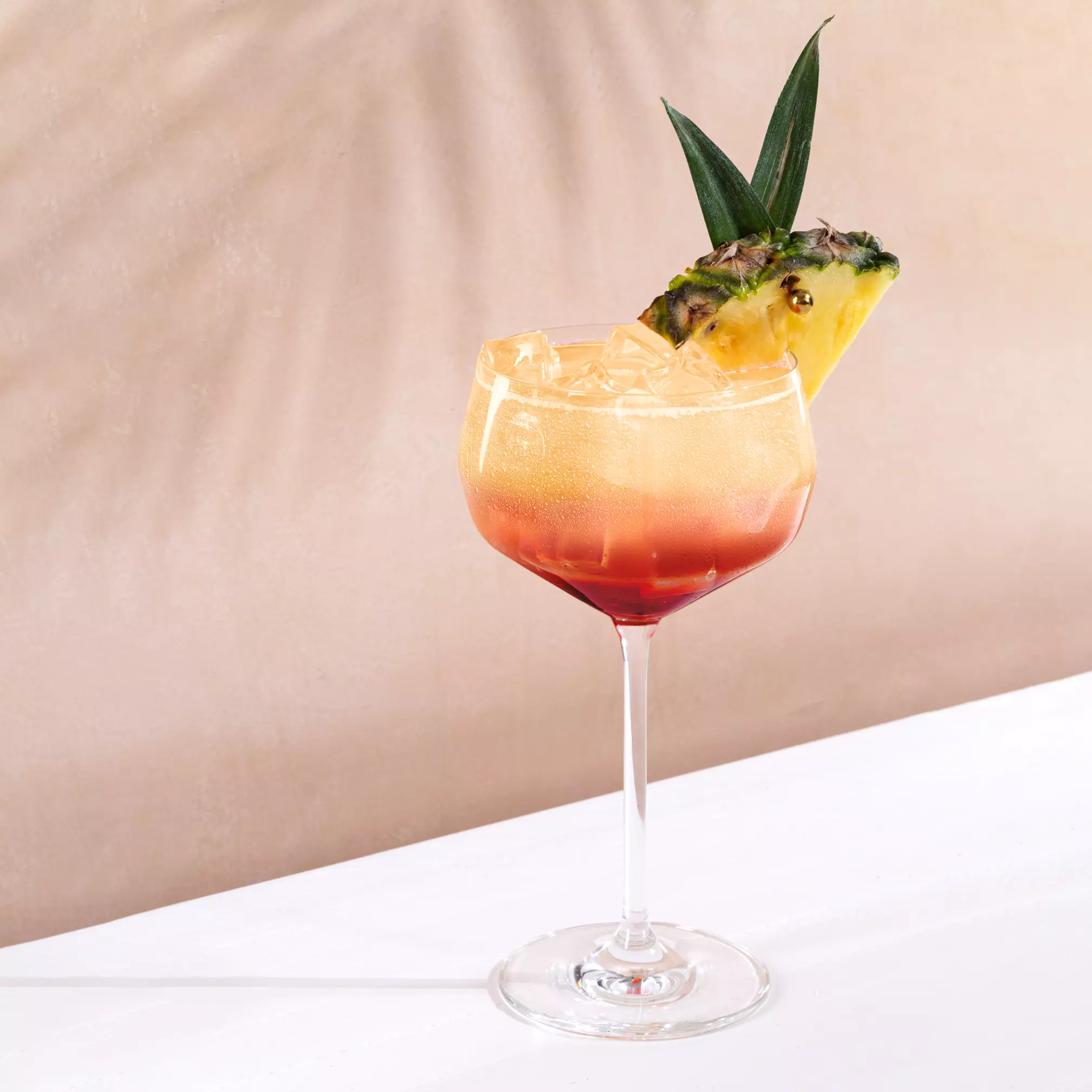 A stemmed glass with a layered cocktail, ice cubes, a slice of pineapple, and a green leaf garnish, set on a white surface against a beige background.