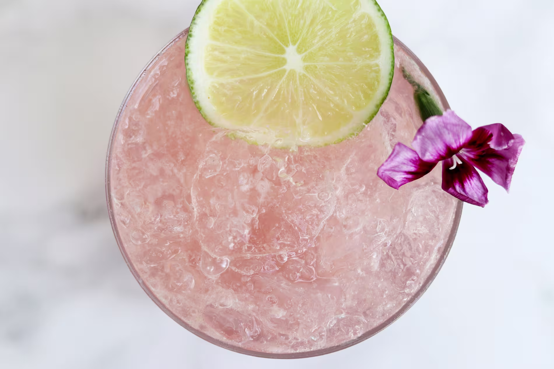 Top view of a pink iced drink garnished with a slice of lime and a purple flower.