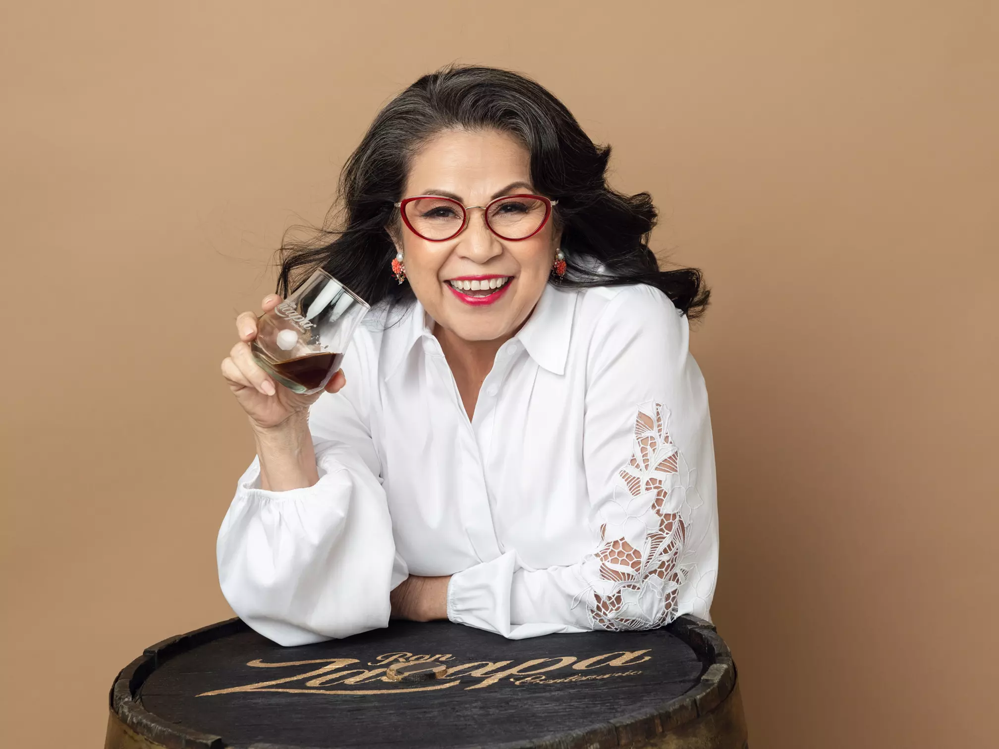 Woman with long dark hair and red glasses smiles while holding a glass of brown liquid, leaning on a barrel with “Ron Zacapa” written on it, against a beige background.