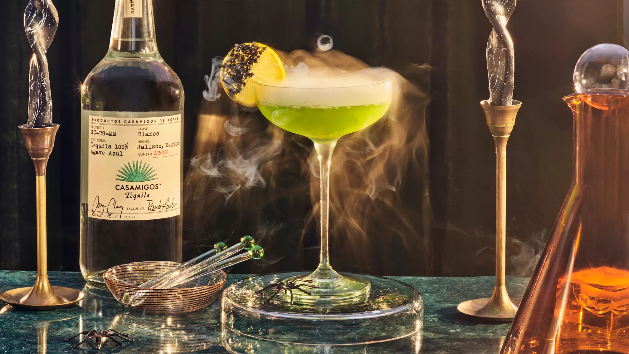 A green cocktail with a smoky effect is served in a coupe glass, garnished with a lemon slice and black salt, surrounded by a tequila bottle, candles, and glass stirrers.