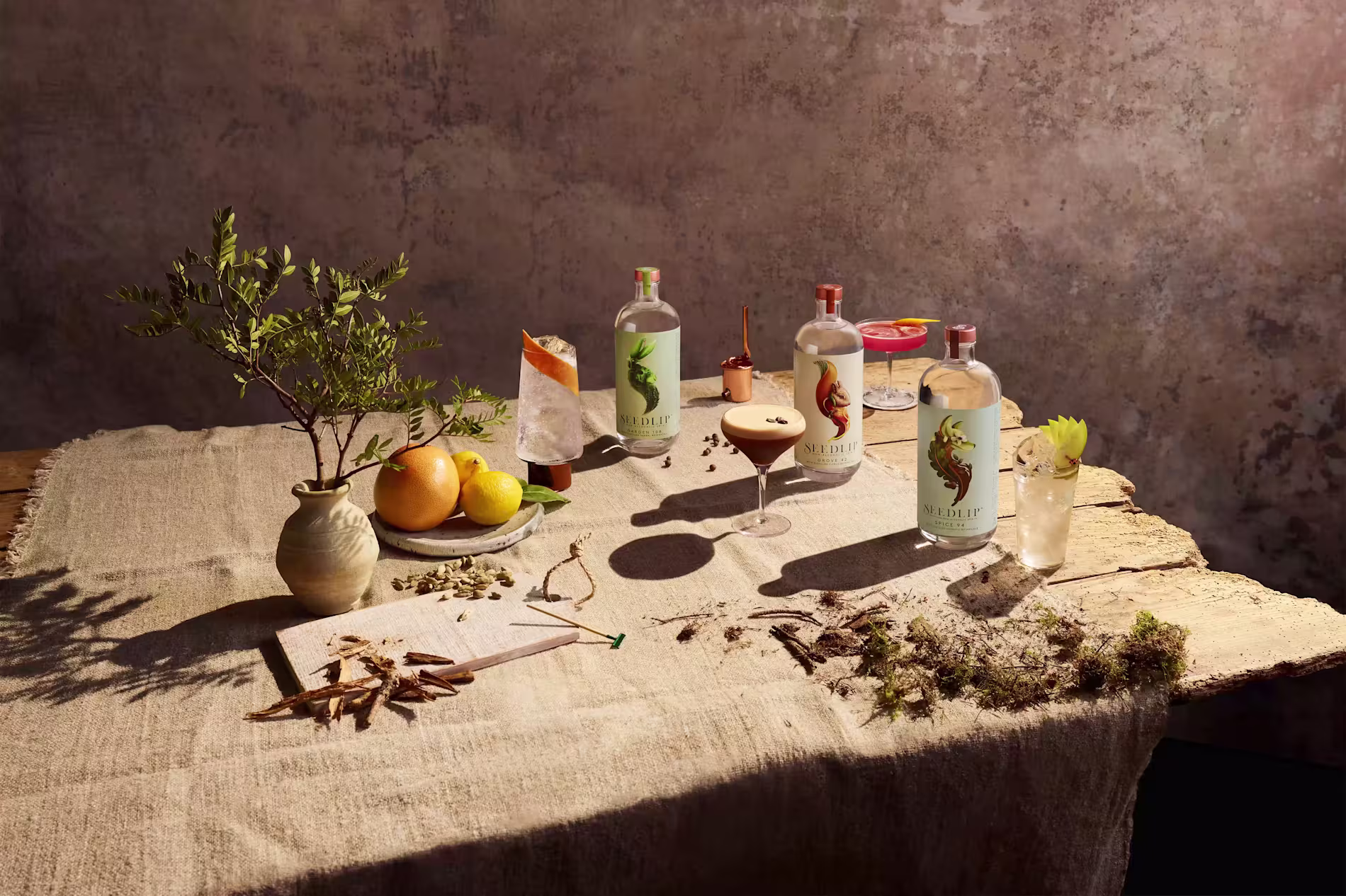 A rustic table displays bottles of gin, cocktails, citrus fruits, a ceramic vase with greenery, and scattered botanicals on a linen cloth.