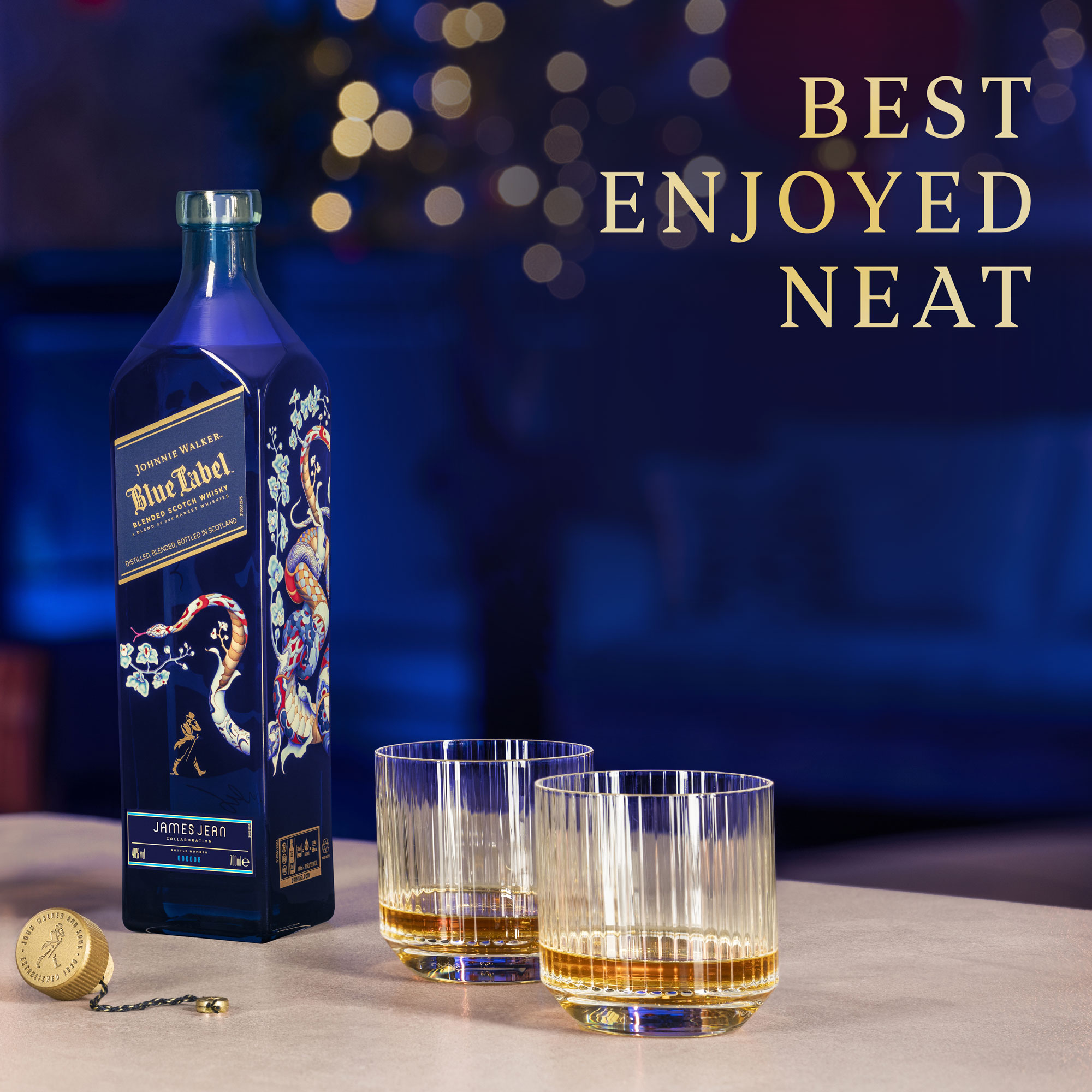 A bottle of Johnnie Walker Blue Label, two glasses with whisky, and a bottle cap sit on a table; text reads Best Enjoyed Neat.