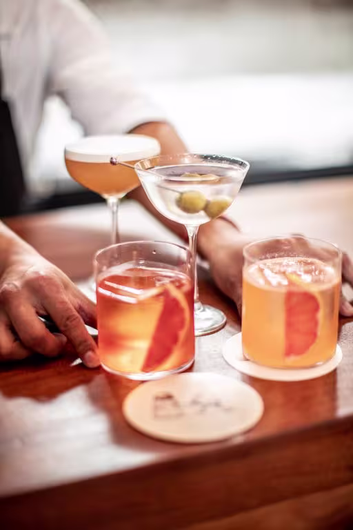 En la imagen se pueden observar cuatro cócteles dispuestos sobre una barra de madera: dos Negroni Rocco en vasos cortos, cada uno adornado con una naranja en su interior, uno colocado sobre un posavasos y el otro apoyado en el costado de un posavasos.
