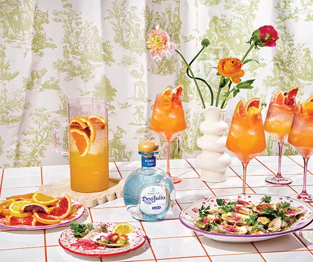 A table with a pitcher and glasses of orange cocktails, a bottle of Don Julio tequila, two plates of food, and a vase with flowers, set against a patterned curtain.