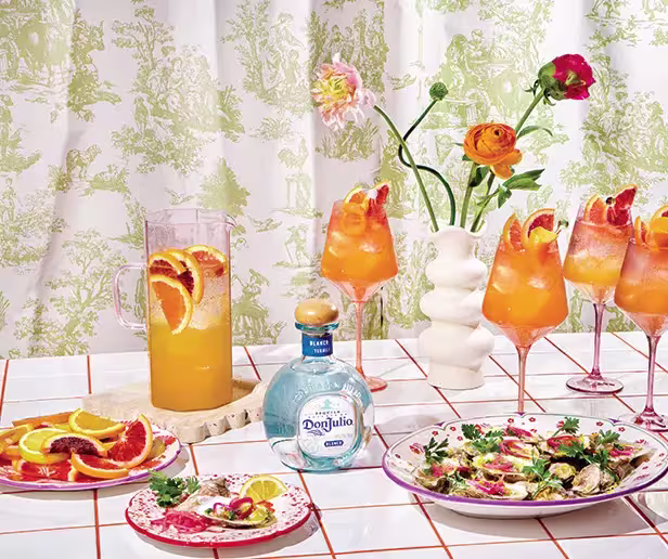 A table with a pitcher and glasses of orange cocktails, a bottle of Don Julio tequila, two plates of food, and a vase with flowers, set against a patterned curtain.