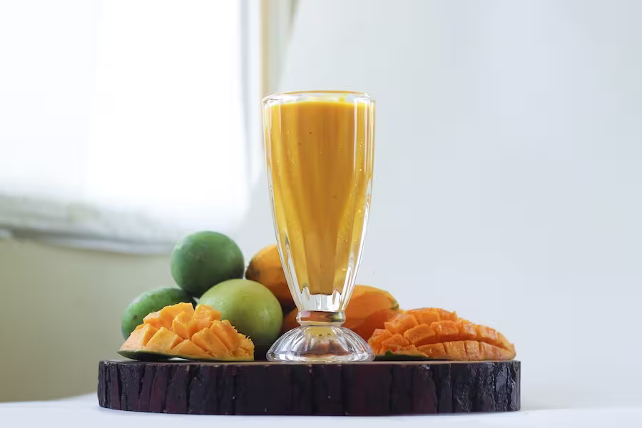 A glass of mango smoothie is placed on a round wooden tray surrounded by whole and sliced mangoes, with a white background.