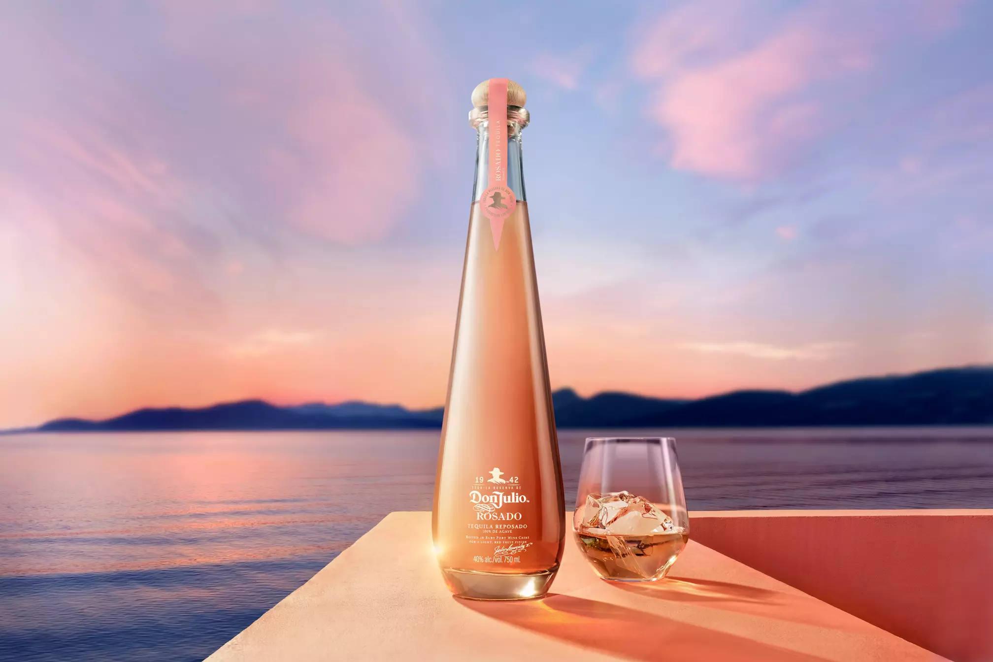 A tall, clear bottle of Don Julio Rosado Tequila and a glass with ice sit on a ledge overlooking a calm body of water at sunset.