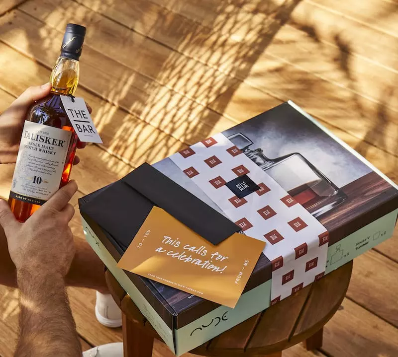 A person holds a bottle of Talisker 10-year-old whisky next to a gift box and a card that reads, This calls for a celebration! on a wooden outdoor table.
