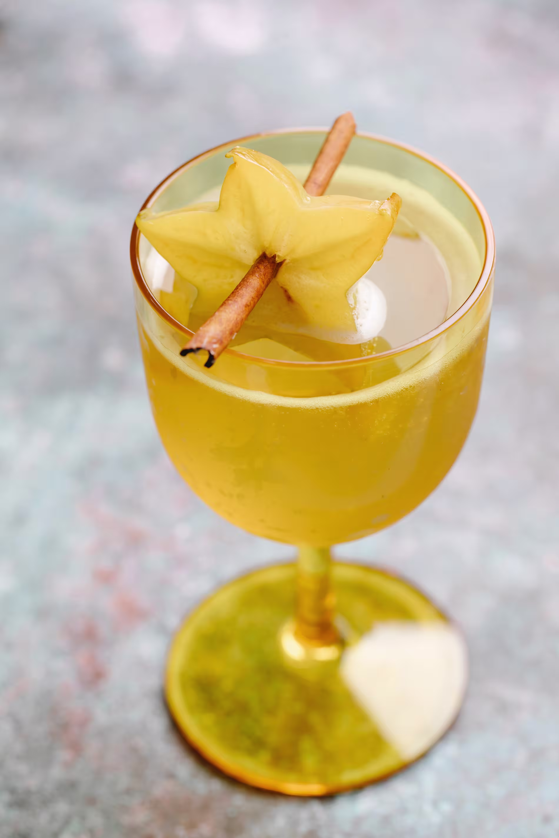 A yellow cocktail in a glass garnished with a starfruit slice and a cinnamon stick.