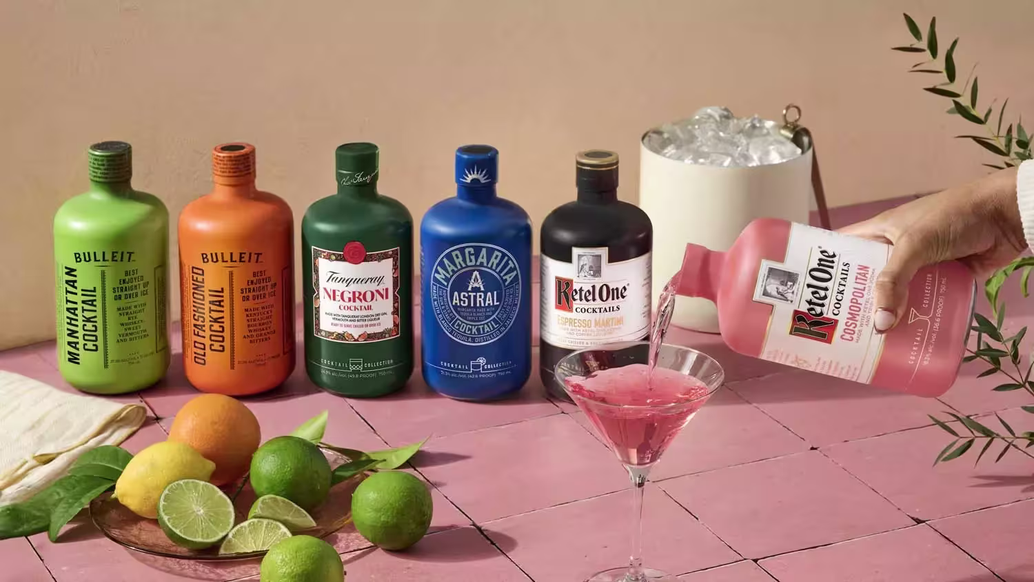Six colorful bottled cocktails and a white ice bucket sit on a pink surface with sliced citrus fruit, while a hand pours pink cocktail into a glass.