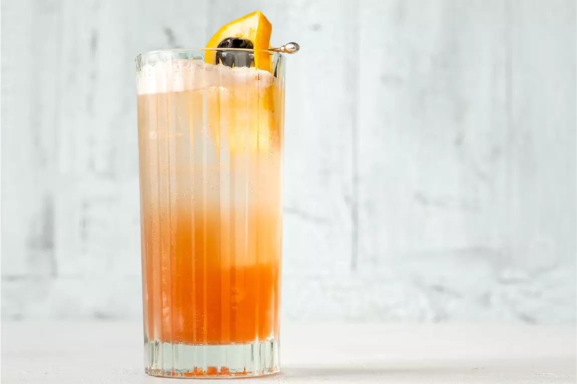 A tall glass of orange and white layered cocktail garnished with an orange slice and cherry on a skewer, set against a light background.