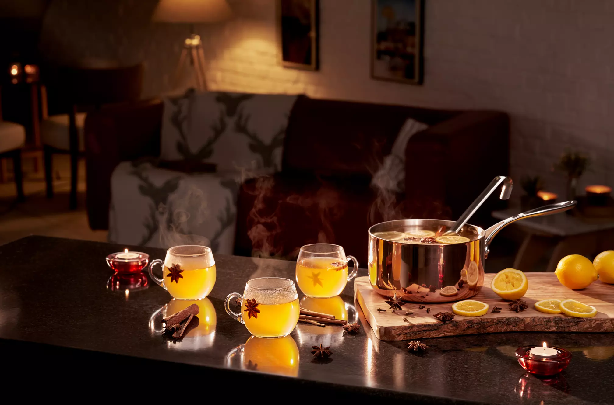 A pot of hot spiced drink sits on a kitchen counter with three filled glass mugs, lemon slices, cinnamon sticks, and star anise, in a warmly lit living room setting.