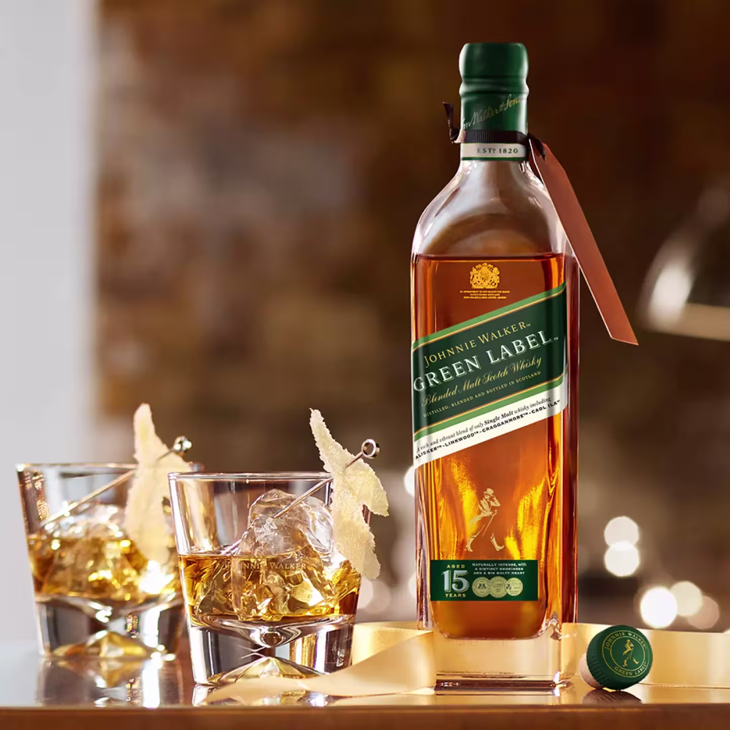 A bottle of Johnnie Walker Green Label Scotch whisky stands on a table next to two glasses with whisky, ice, and decorative slices. The bottle cap lies nearby.