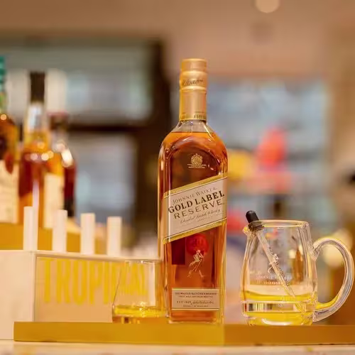 A bottle of Johnnie Walker Gold Label Reserve whisky is displayed next to a glass mug, a glass, and a dropper in a well-lit setting.