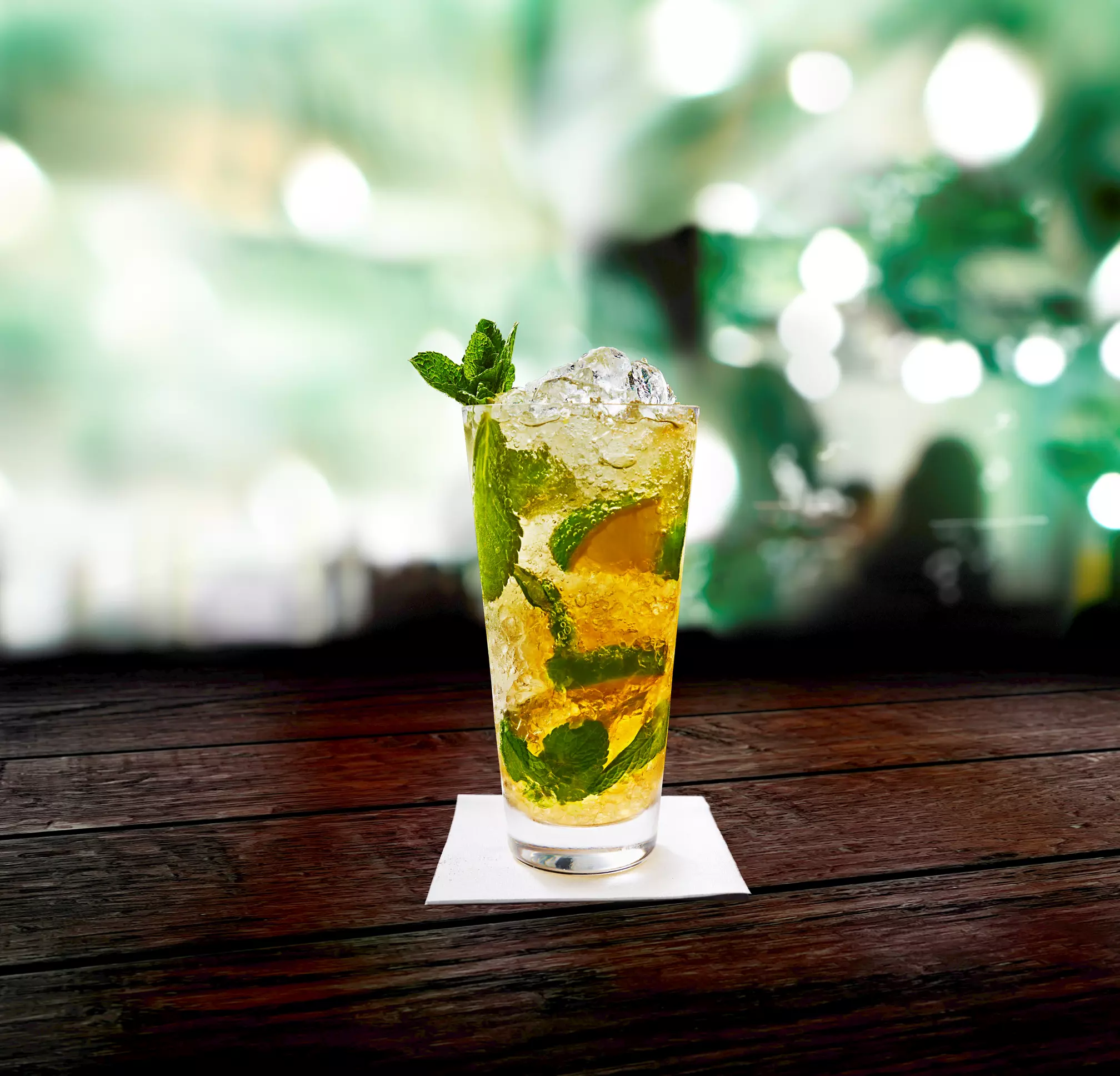 A glass of iced cocktail with mint leaves and lime, garnished with mint, sits on a wooden table with a blurred green and white background.
