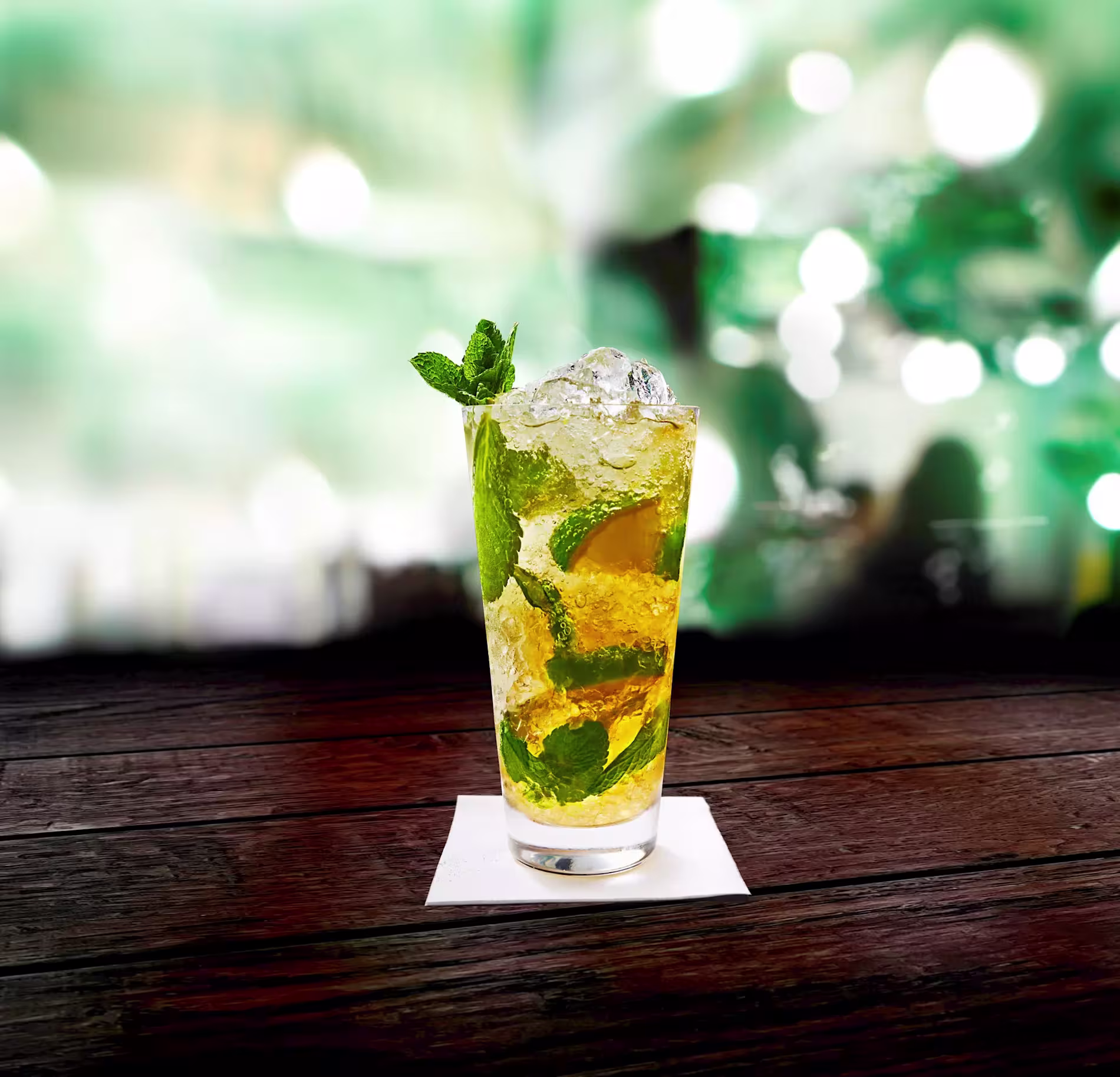 A glass of iced cocktail with mint leaves and lime, garnished with mint, sits on a wooden table with a blurred green and white background.