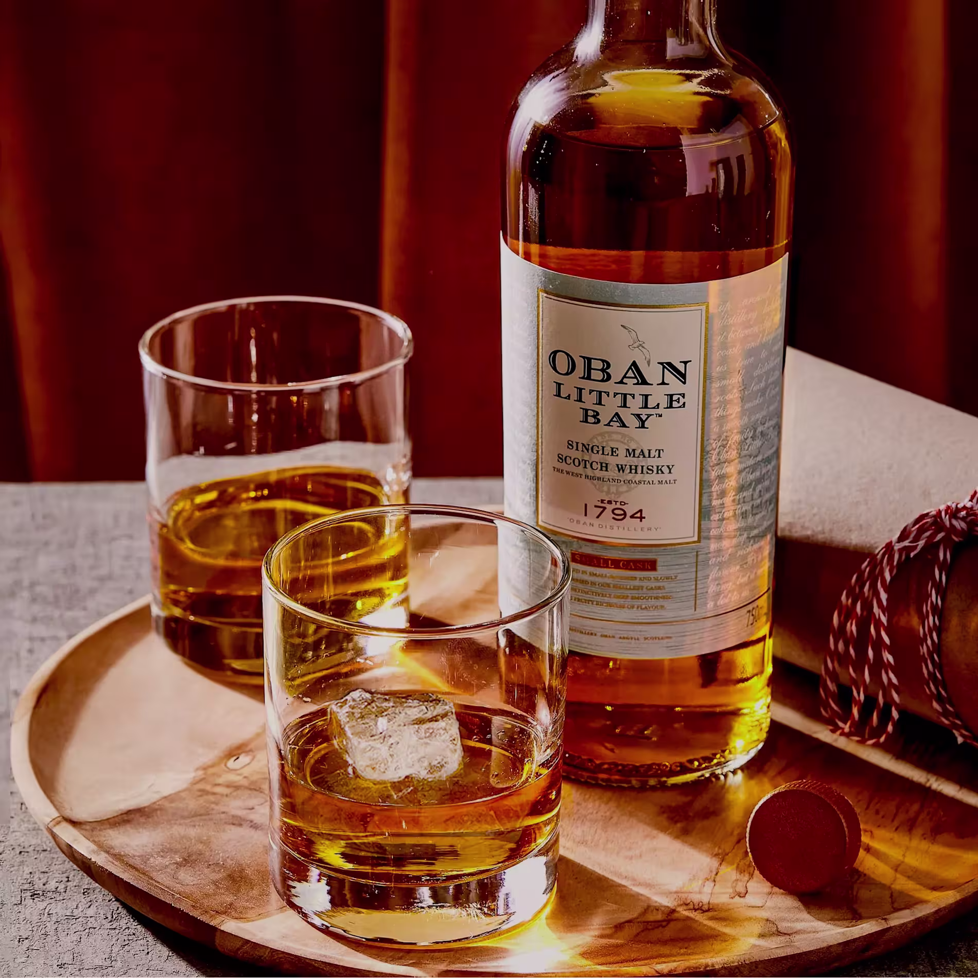 A bottle of Oban Little Bay single malt scotch whisky on a wooden tray with two glasses of whisky, one with ice.